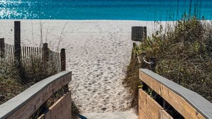 Un paseo marítimo de madera que conduce a una playa arenosa, con un cielo azul claro y el océano al fondo.
