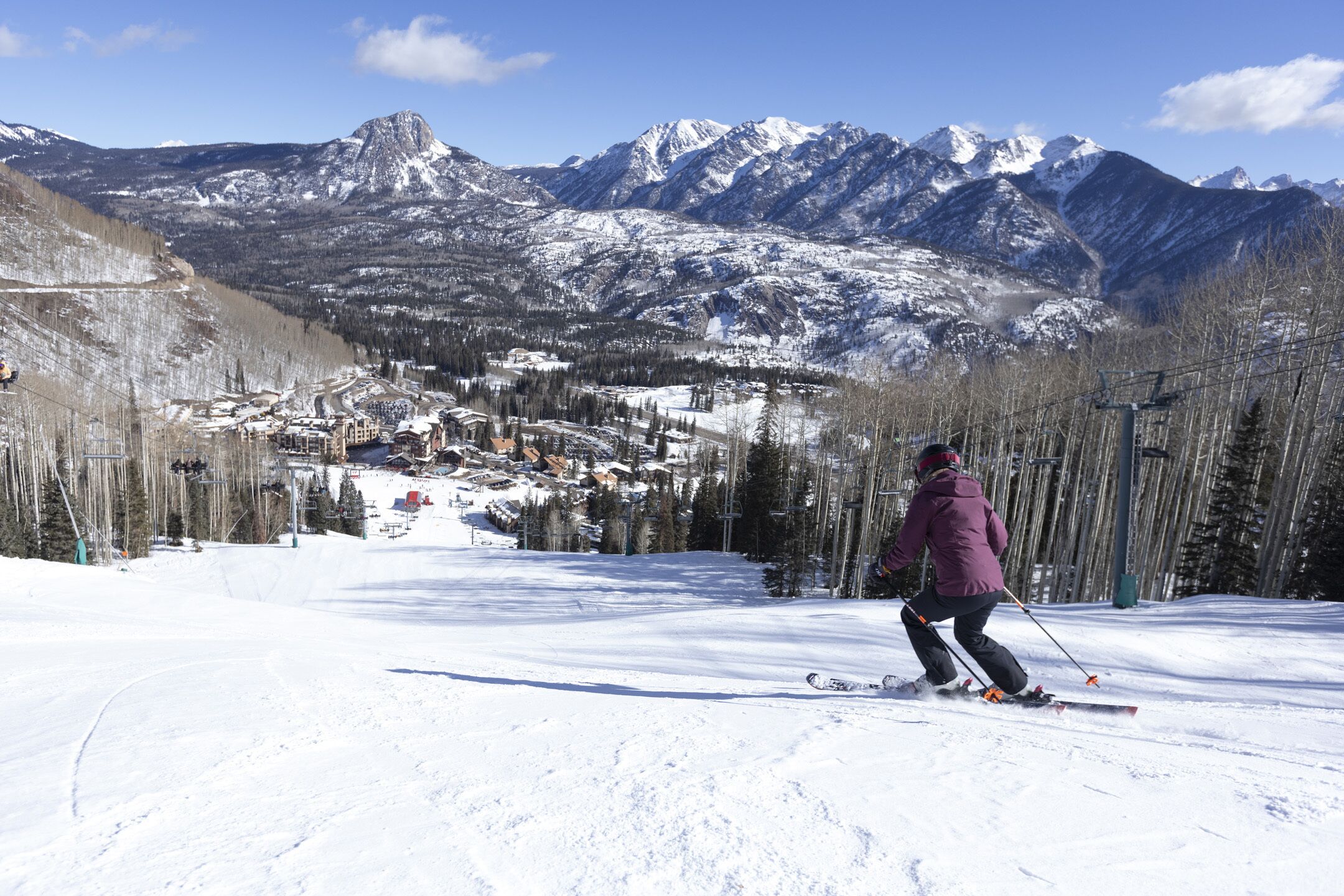 A ski slope with a person skiing down, mountains in the background, and a small town at the base.