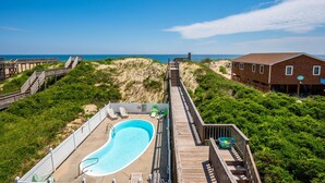 A wooden deck leading to a swimming pool with lounge chairs, surrounded by greenery and a beach view.