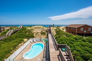 A wooden deck leading to a swimming pool with lounge chairs, surrounded by greenery and a beach view.