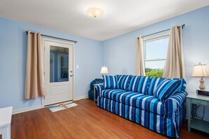 A room with a blue-striped sofa, a door with beige curtains, a window with beige curtains, a small table with a lamp, and a digital clock. - Absolute Searenity- Oceanfront, Pet-Friendly, Private Pool and Hot Tub (Nags Head)