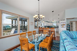 A spacious living area with a glass dining table surrounded by wooden chairs, a blue-striped sofa, and a kitchen in the background. - Absolute Searenity- Oceanfront, Pet-Friendly, Private Pool and Hot Tub (Nags Head)