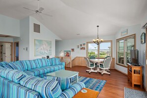 A living room with a striped sofa, a dining table with chairs, a ceiling fan, and artwork on the walls. - Absolute Searenity- Oceanfront, Pet-Friendly, Private Pool and Hot Tub (Nags Head)
