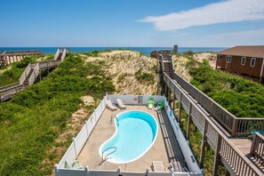 Une piscine dotée d’une terrasse en bois et de chaises longues, entourée d’une clôture en bois et de végétation, et offrant une vue sur l’océan et les dunes.