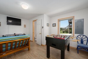 A room with a foosball table, a bed with striped bedding, a television on the wall, and a window with a view of a wooden deck. - Absolute Searenity- Oceanfront, Pet-Friendly, Private Pool and Hot Tub (Nags Head)