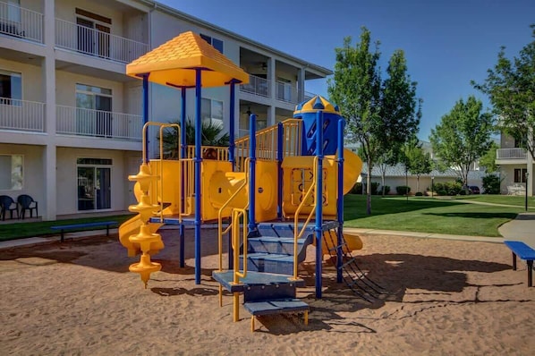 Un terrain de jeux équipé d’un toboggan et de structures d’escalade, situé devant un immeuble à plusieurs étages doté de balcons.
