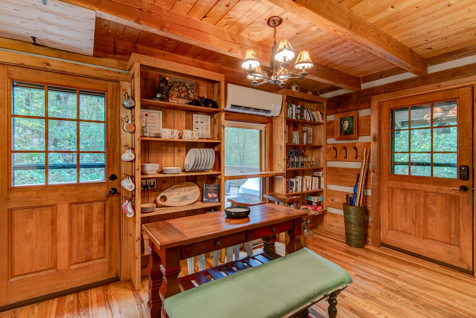 Un interior de madera con una mesa de comedor, bancos, estanterías con libros y utensilios de cocina, y una araña.