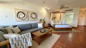 Uma sala de estar com sofá, mesa de centro e cama com dossel. Há duas obras de arte penduradas na parede e um ventilador de teto.