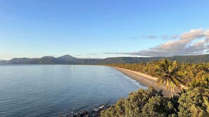 Paisagem costeira com praia, palmeiras e montanhas ao fundo.