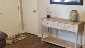 Ein Zimmer mit Holzboden, einer weißen Tür, einem Konsolentisch mit Schubladen, einer Vase und einem Spiegel an der Wand.