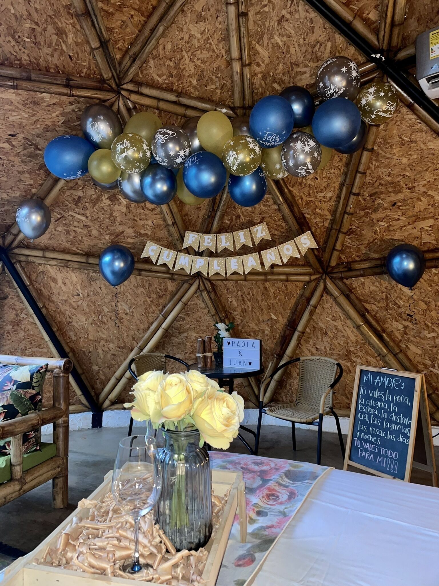 A room with a geometric wooden ceiling, a table with a floral tablecloth, a vase of yellow roses, and a sign with a message.