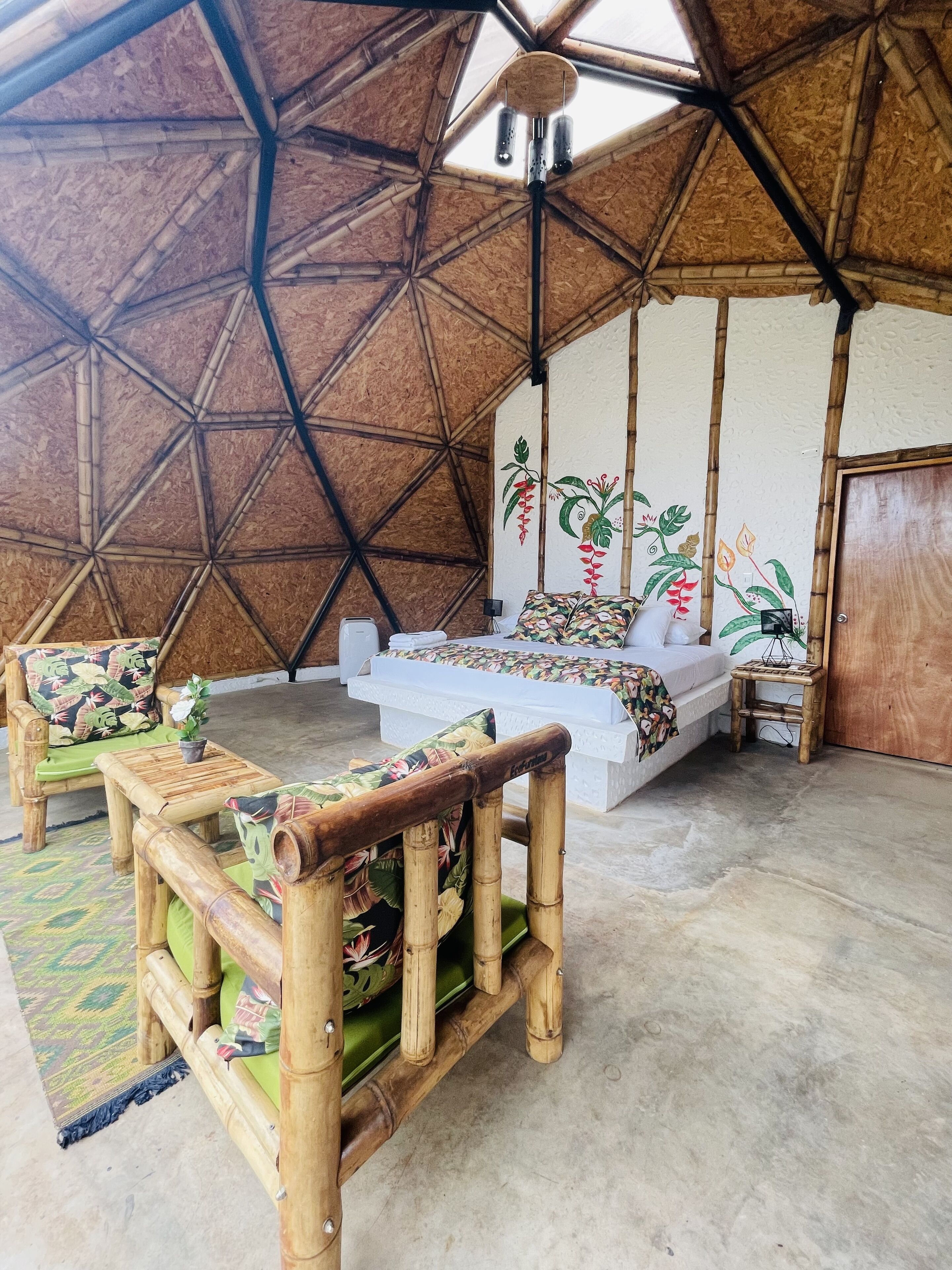Quarto com teto geométrico de bambu, cama com colcha florida e uma mesinha com uma planta.