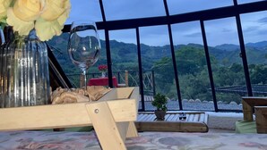 A room with a large window offering a view of green hills. There is a wooden table with a vase of flowers and a glass. - Ecofuratena Glamping (Utica)
