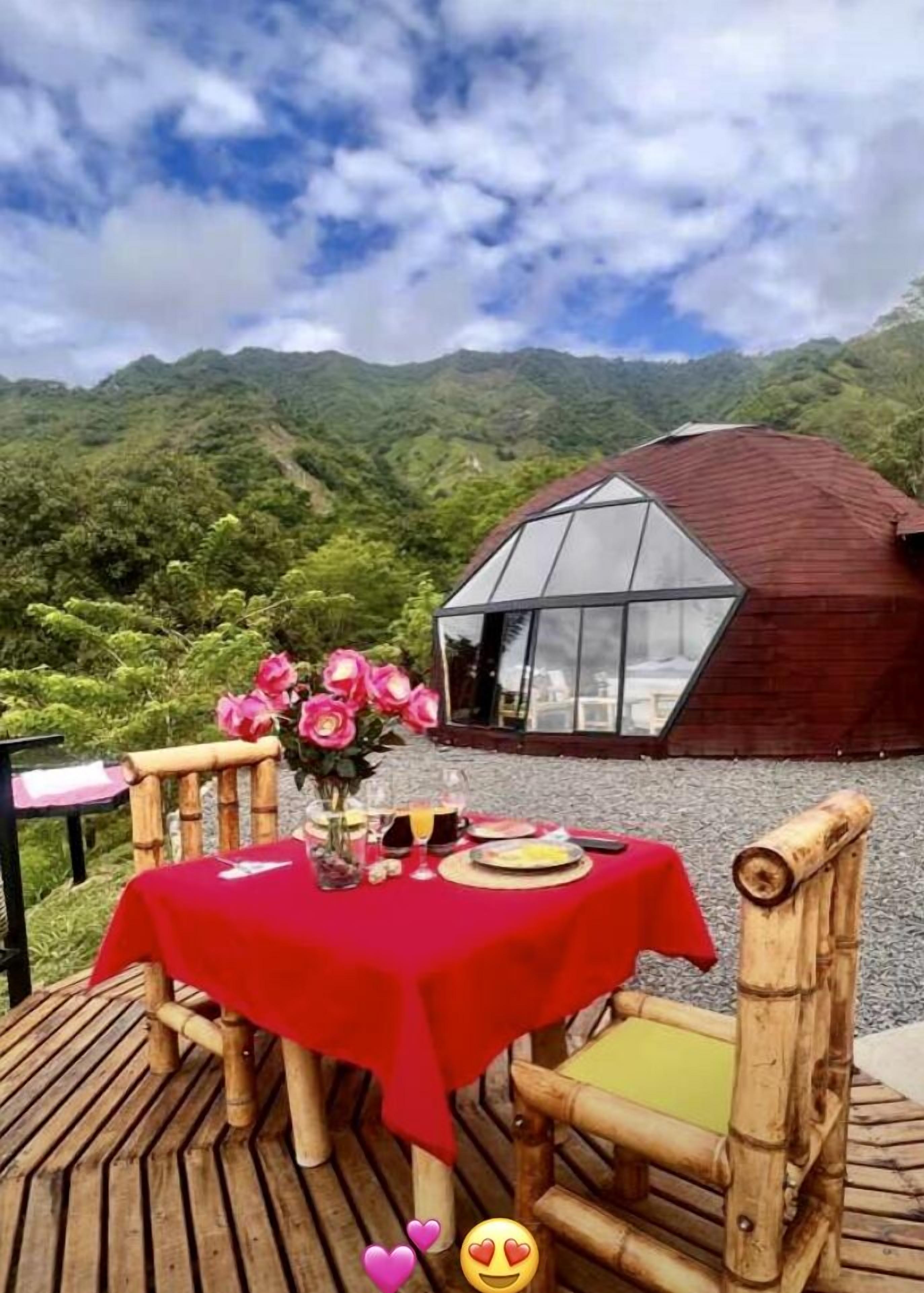 A table set for two with a red tablecloth, a vase of pink flowers, and a view of a mountain landscape.