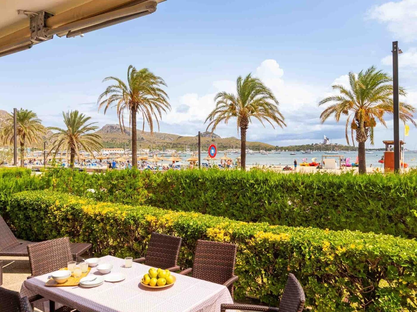 A table set with plates, glasses, and a bowl of lemons, overlooking a beach with palm trees and boats.
