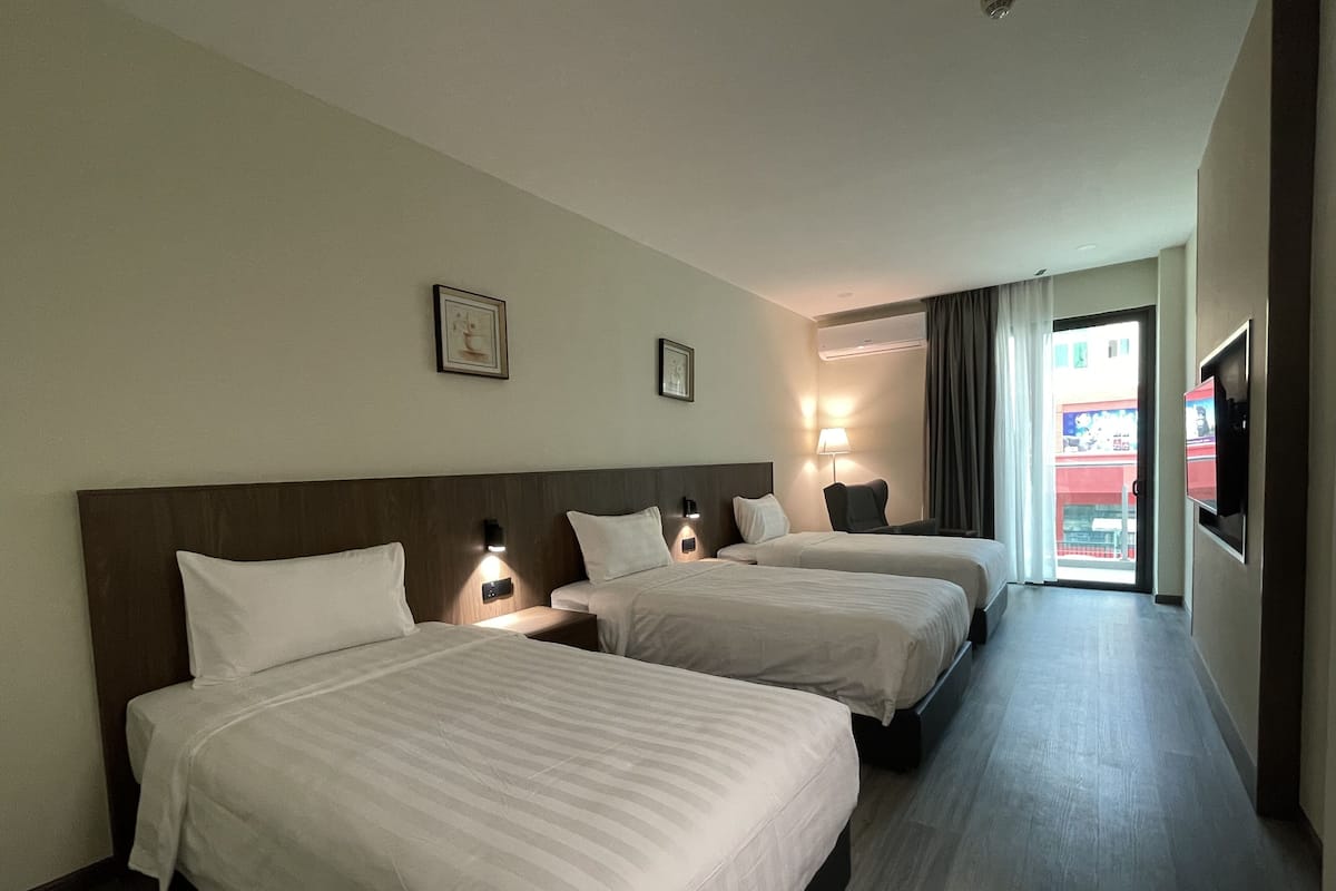 Ein Hotelzimmer mit drei Betten, einem Fernseher, einer Lampe und einem Fenster mit Blick auf ein Gebäude.