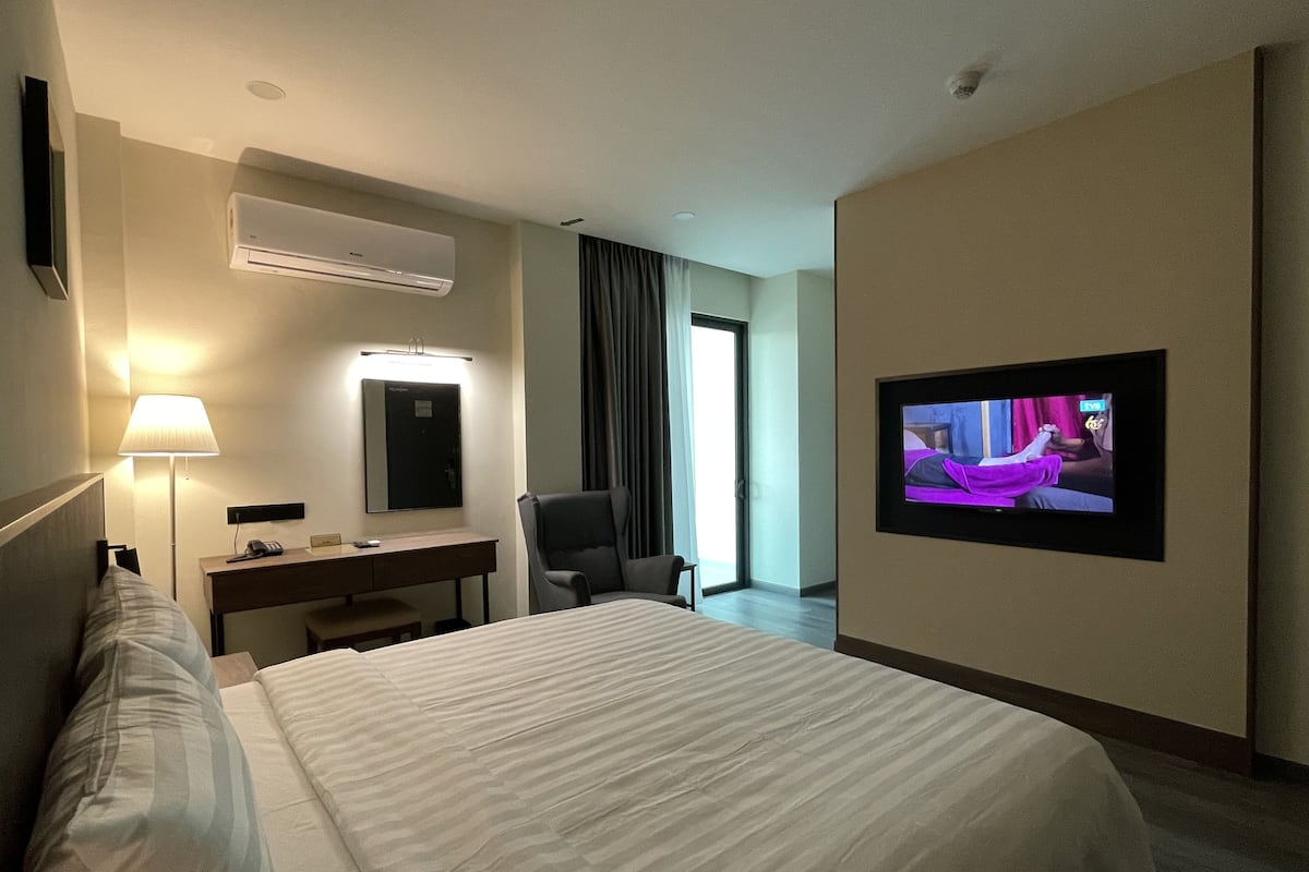 Ein Hotelzimmer mit einem großen Bett, einem Schreibtisch mit Lampe, einem Sessel, einem Fernseher und einer Klimaanlage.