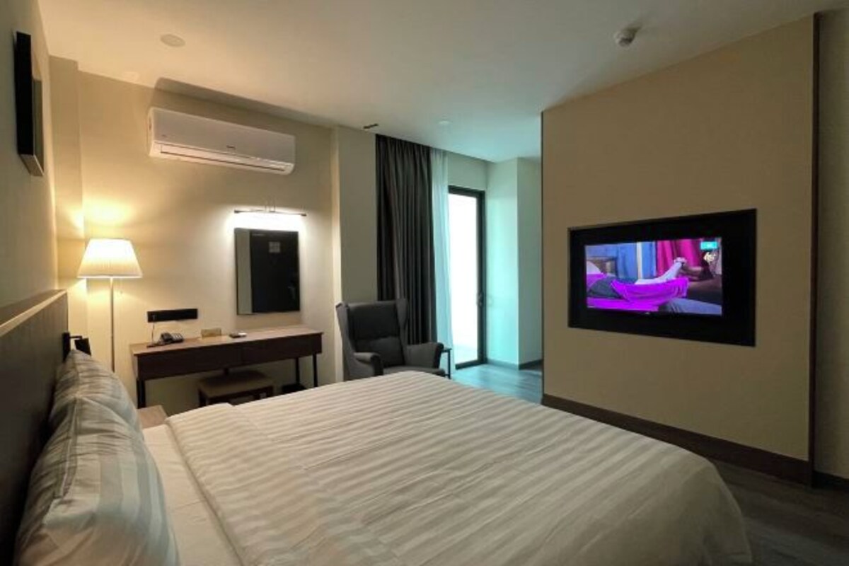 A hotel room with a bed, a desk, a chair, a TV, and an air conditioning unit.