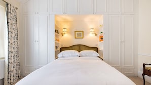 Una cama bien hecha en una habitación con paredes blancas y un cabecero de madera. Hay luces fijadas a la pared y un cuadro enmarcado sobre la cama.
