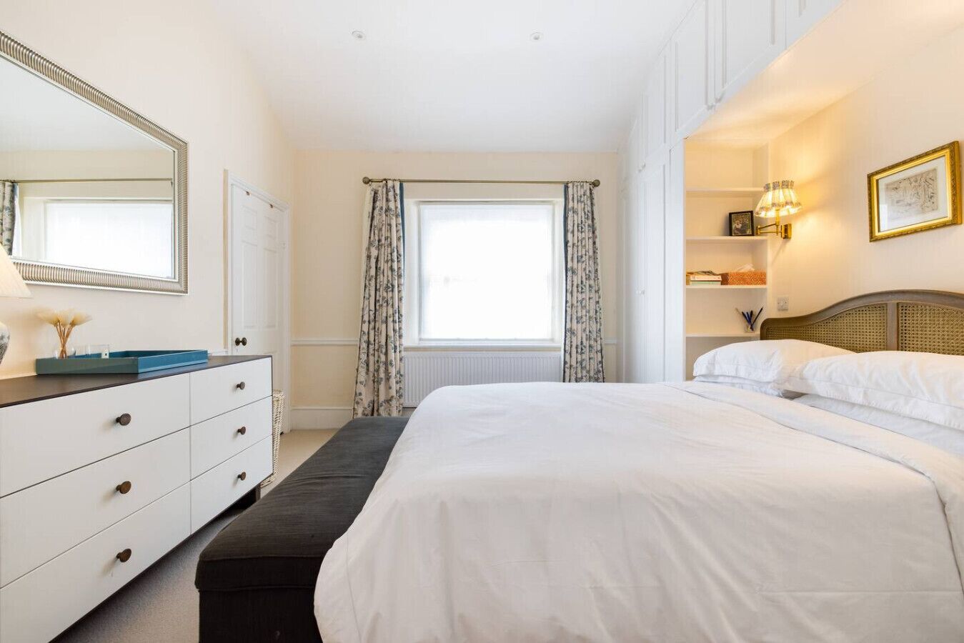 A bedroom with a large bed, a dresser, a mirror, a window with curtains, and a bedside table with a lamp.