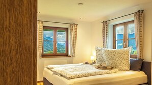 A bedroom with a bed, two windows with curtains, a nightstand, and a view of mountains.