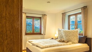 Een slaapkamer met een bed, twee ramen met gordijnen, een bedlamp en uitzicht op bergen.