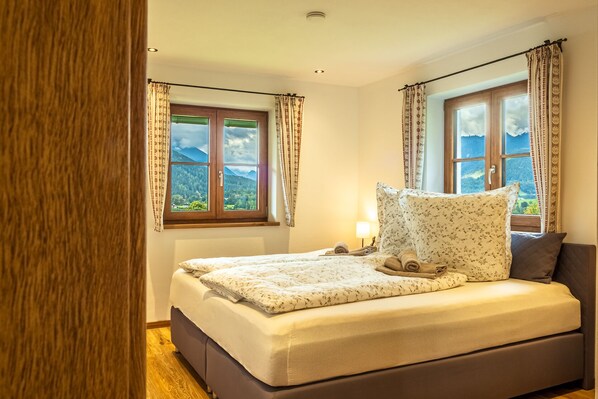 A bedroom with a bed, two windows with curtains, a bedside lamp, and a view of mountains.