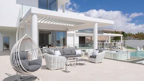 Un espace de vie extérieur moderne, doté d’une piscine, de meubles en rotin blanc et d’un fauteuil suspendu.