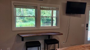 Ein Zimmer mit einer hölzernen Bank, zwei schwarzen Barhocker, einem montierten Fernseher und einem Fenster mit Blick auf Grünflächen.