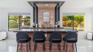 A modern kitchen with a large island, bar stools, and large windows with a view of greenery. - Multimillion $ ResortVilla+Waterslide+Pickleball+ (Scottsdale)
