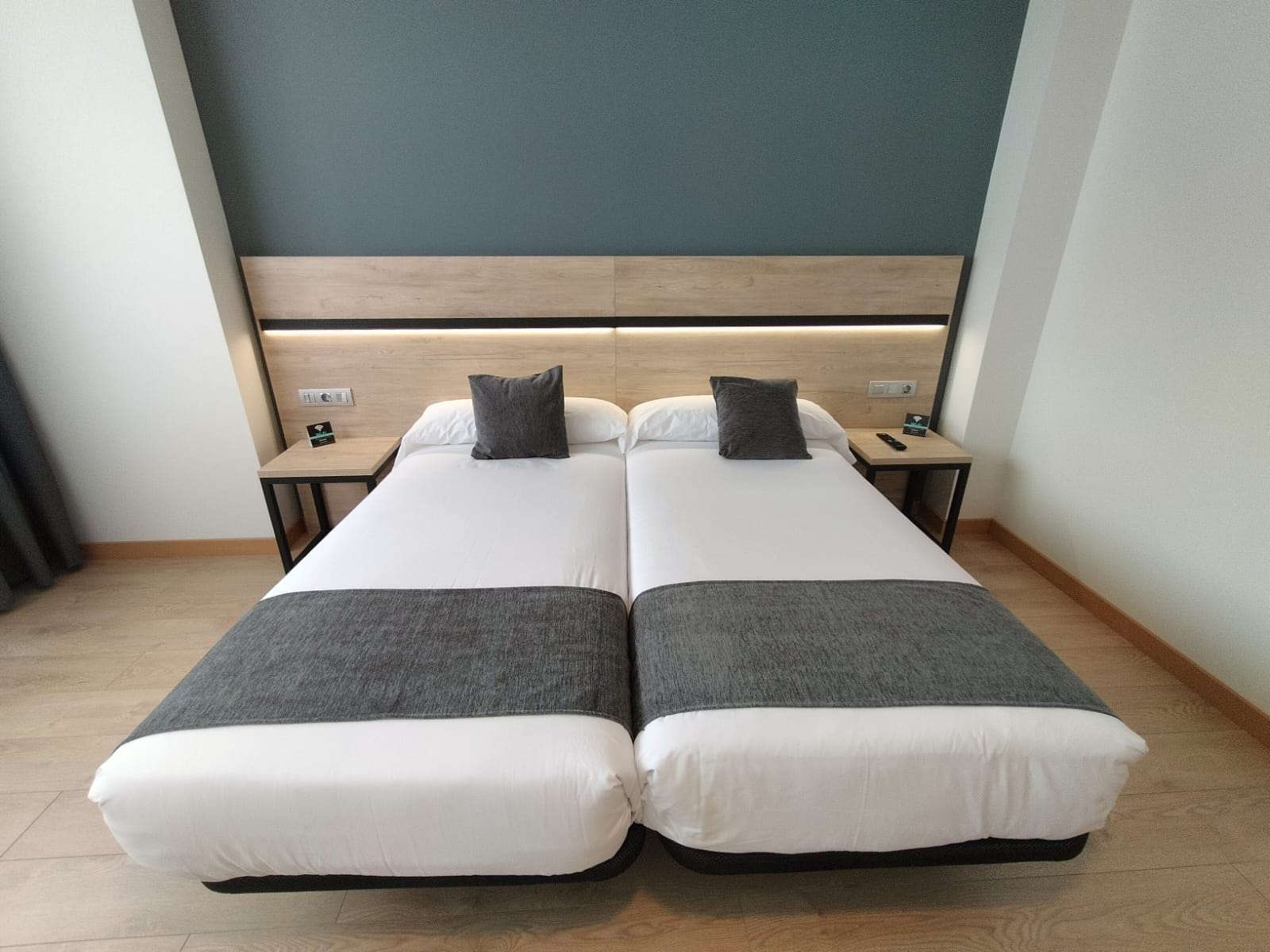 Habitación doble con dos camas, cada una con almohada y manta. Hay mesitas de noche con teléfono y control remoto. La habitación tiene un cabecero de madera y una pared lisa.
