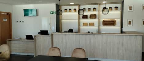 A hotel reception area with a wooden reception desk, a television mounted above it, and a wall with shelves displaying wicker baskets and potted plants.