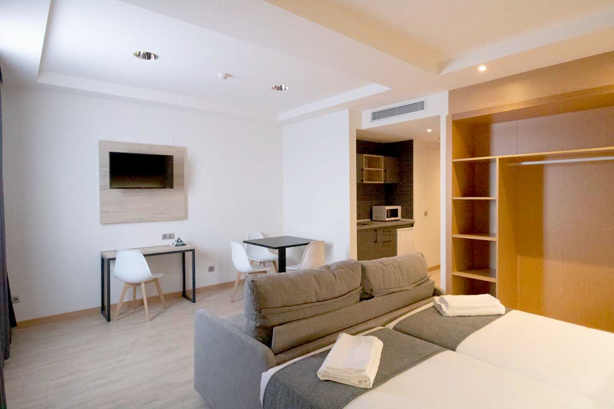 A modern hotel room with a sofa bed, a desk, a flat-screen TV mounted on the wall, and a walk-in closet.