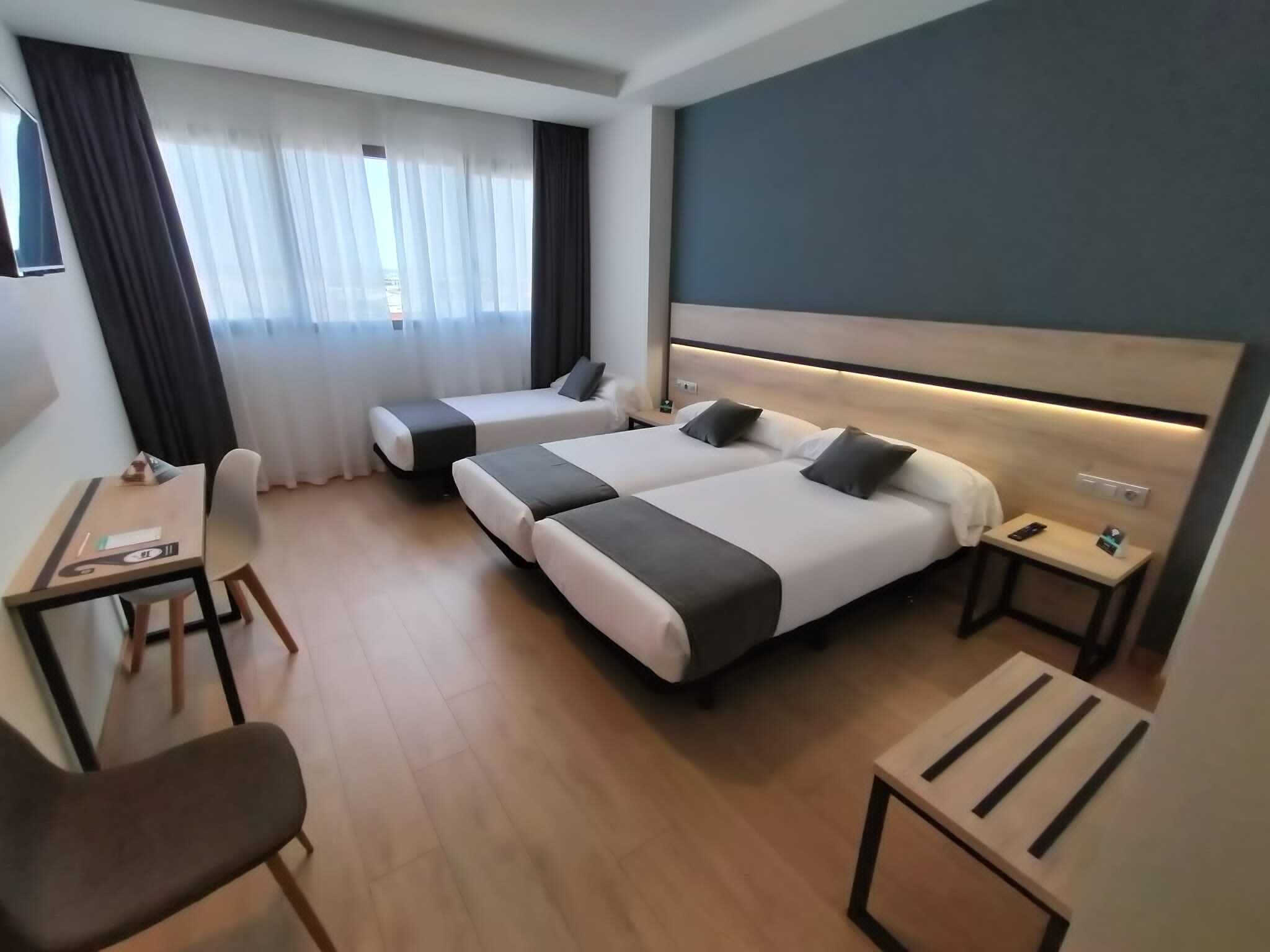 A hotel room with two single beds, a desk with a chair, a small table, and a nightstand with a lamp.