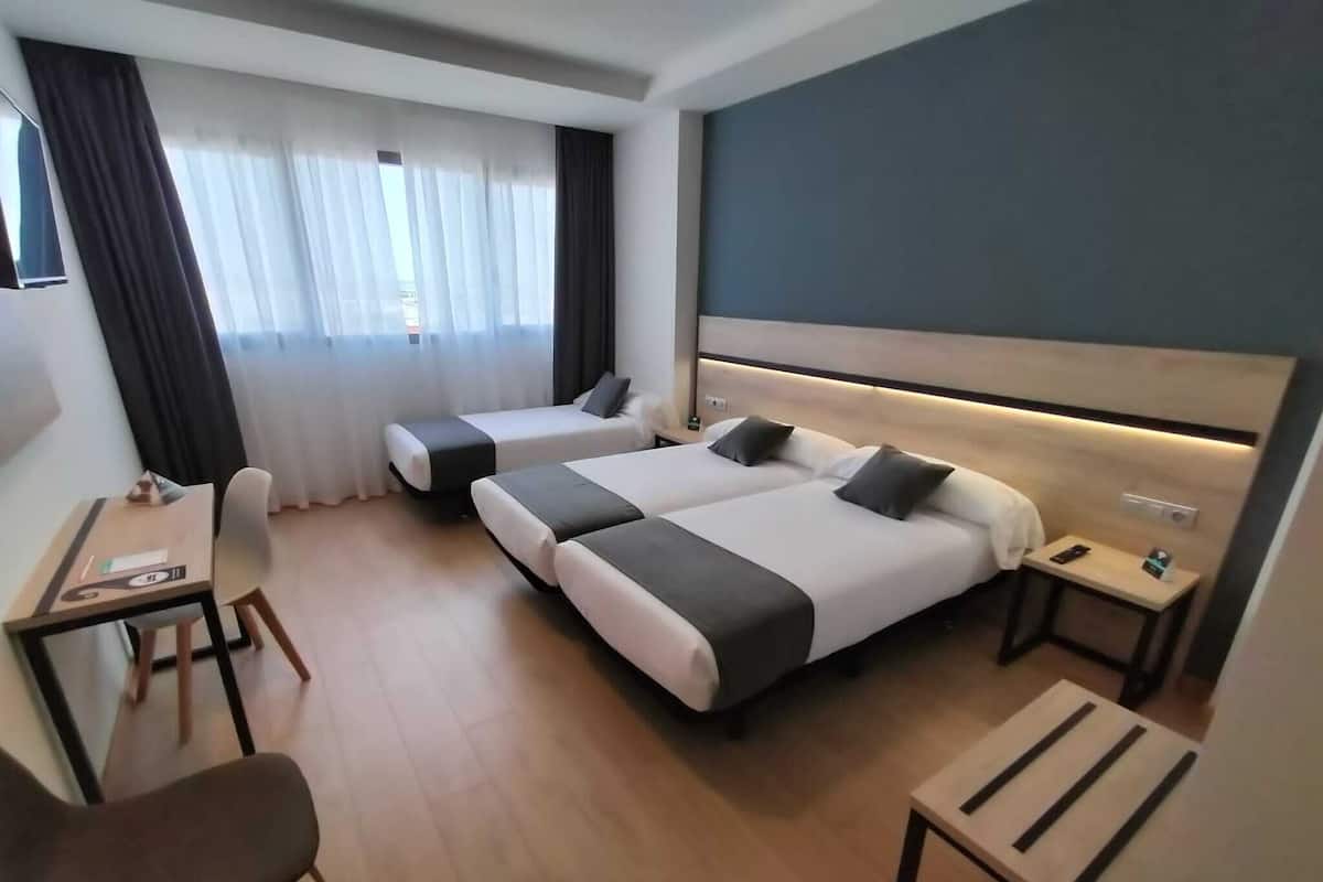Quarto de hotel com duas camas de solteiro, uma escrivaninha com cadeira, uma mesinha e uma mesa de cabeceira com abajur.