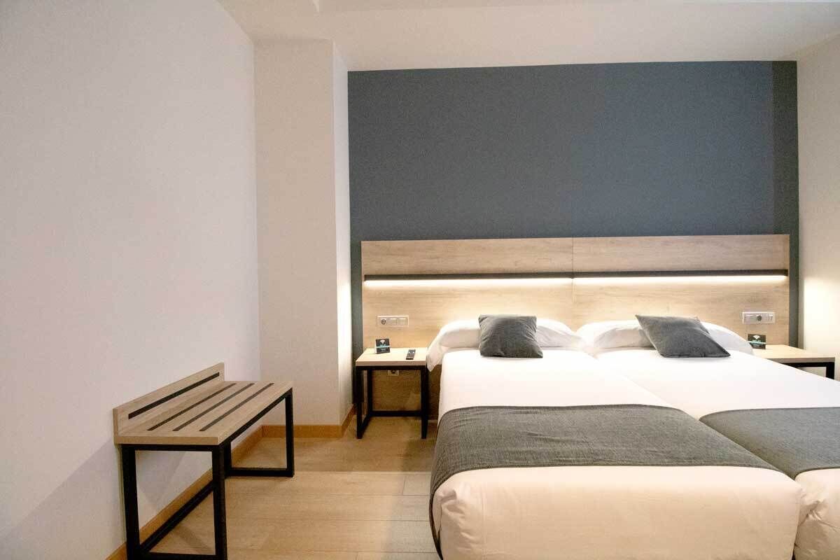 A hotel room with a large bed, a bedside table, a bench, and a headboard with built-in lighting.