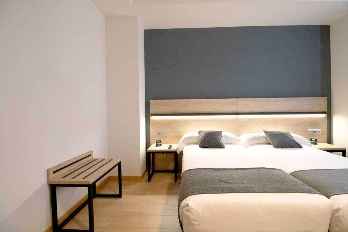 Quarto de hotel com uma cama grande, uma mesa de cabeceira, um banco e cabeceira com iluminação embutida.