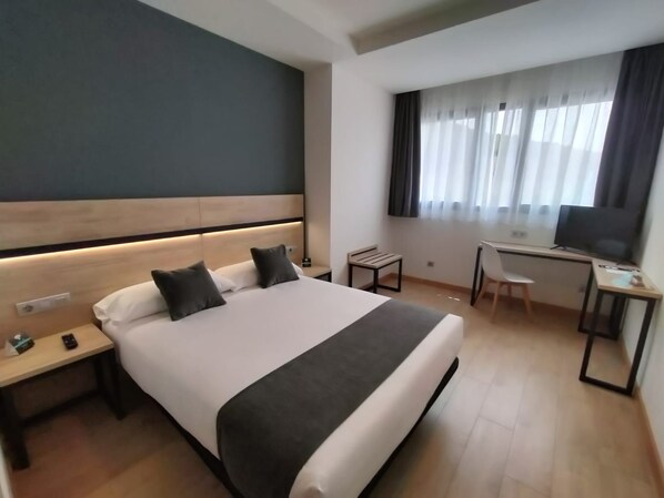 Una habitación de hotel con cama, mesitas de noche, un escritorio con silla, televisor y ventana con cortinas.