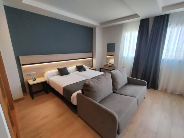 A hotel room with a bed, a sofa, a nightstand, and a window with curtains.