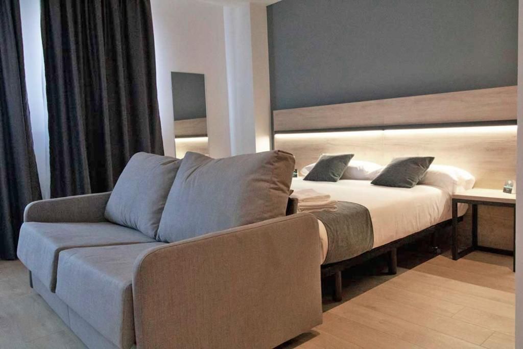 Una habitación de hotel moderna con sofá, cama, mesita de noche y cabecera con iluminación integrada.