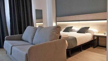Una habitación de hotel moderna con sofá, cama, mesita de noche y cabecera con iluminación integrada.