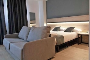 A modern hotel room with a sofa, a bed, a nightstand, and a headboard with built-in lighting. - Alda Valladolid Sur (Valladolid)