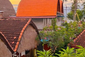 Une maison traditionnelle à toit de chaume, surmontée d’un toit de tuiles rouges, et entourée d’une végétation luxuriante.