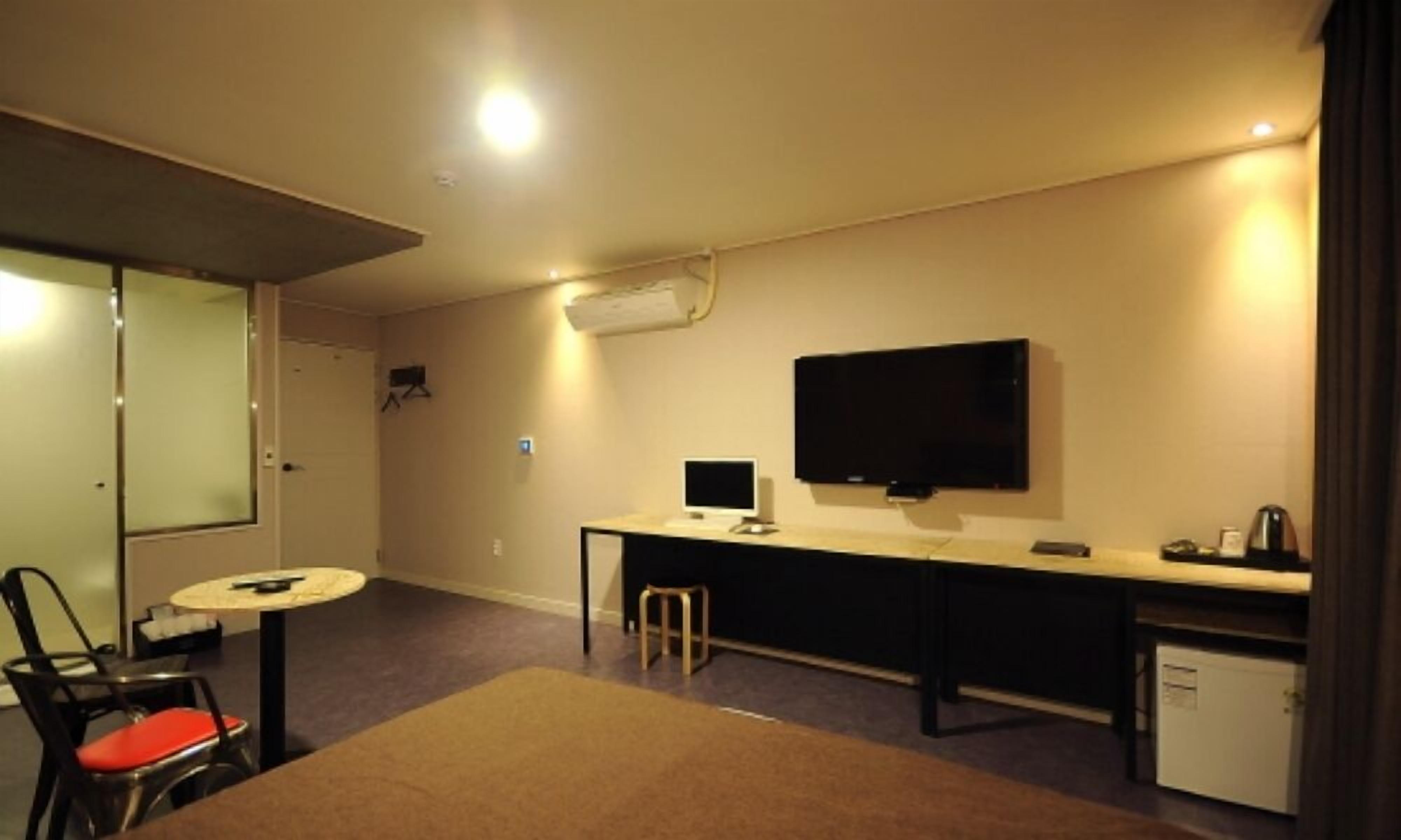 A hotel room with a bed, a desk with a computer, a TV, a small kitchenette, and a chair with a red cushion.