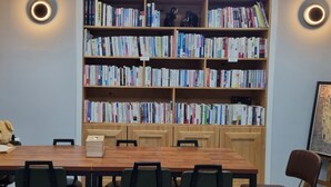 A room with a wooden table, chairs, a bookshelf filled with books, a teddy bear, and a framed picture on the wall. - HOTEL 385 (Changwon)