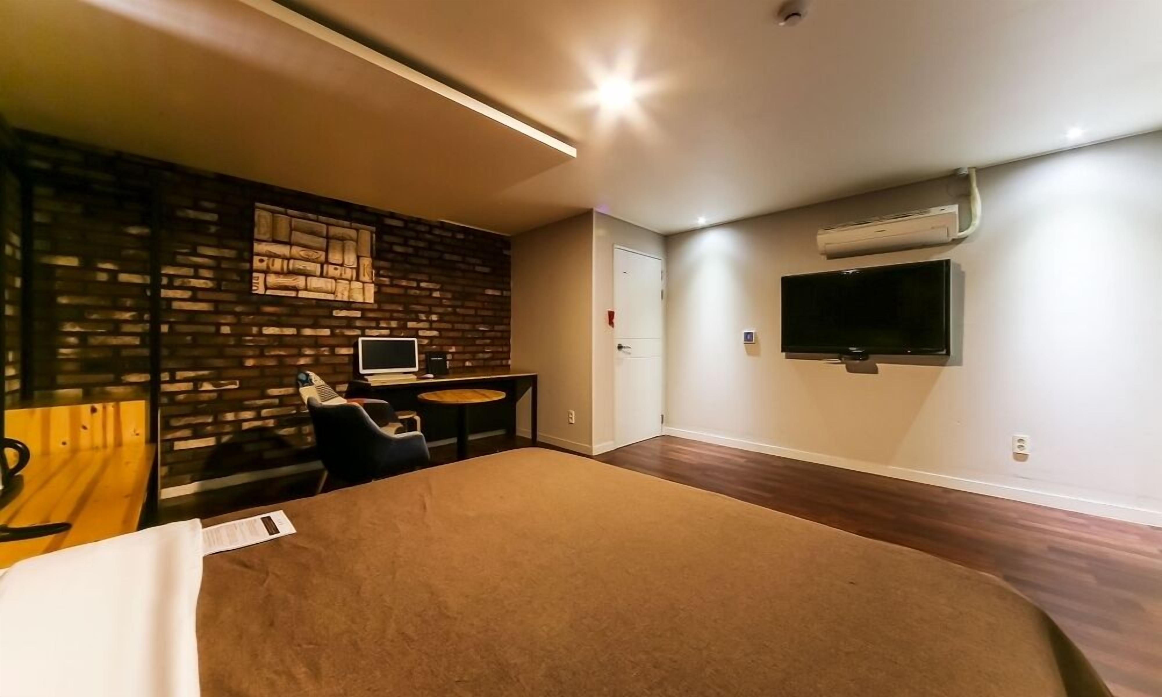 Una habitación de hotel moderna con pared de ladrillo decorativa, un televisor de pantalla plana montado en la pared, un escritorio con computadora y una cama grande con cubierta marrón.