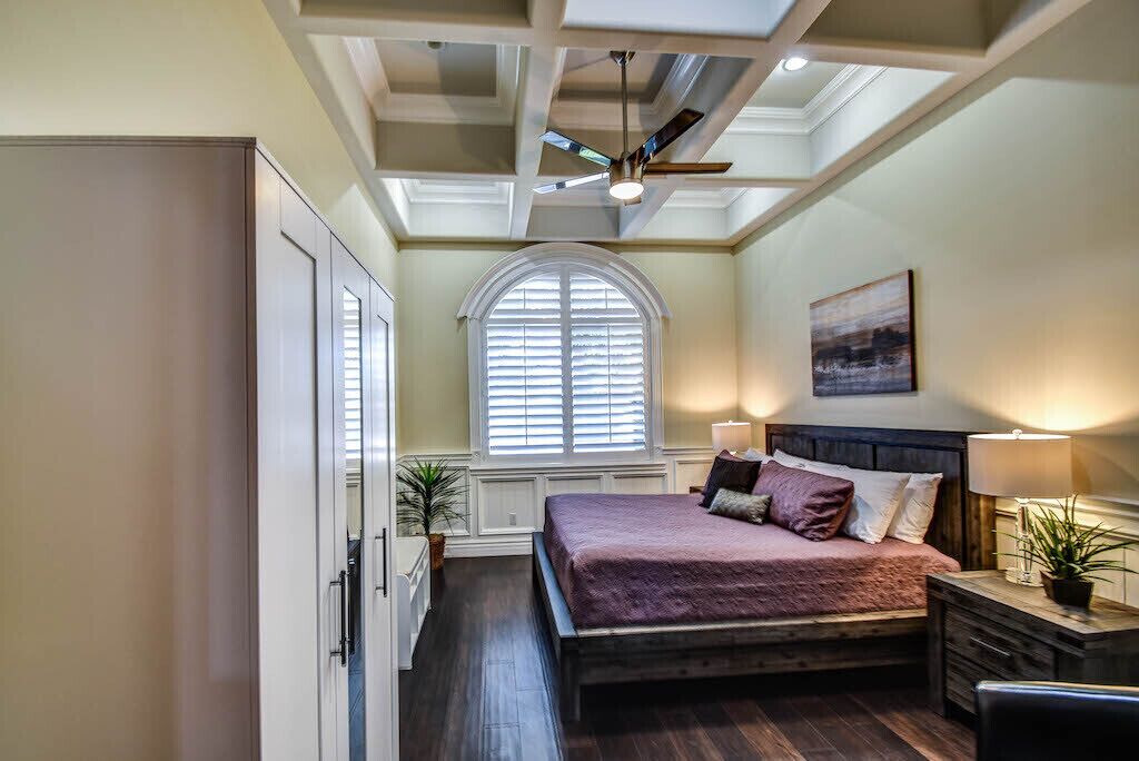 Une chambre à coucher avec un lit, un ventilateur de plafond, une fenêtre avec des stores et un tableau accroché au mur.