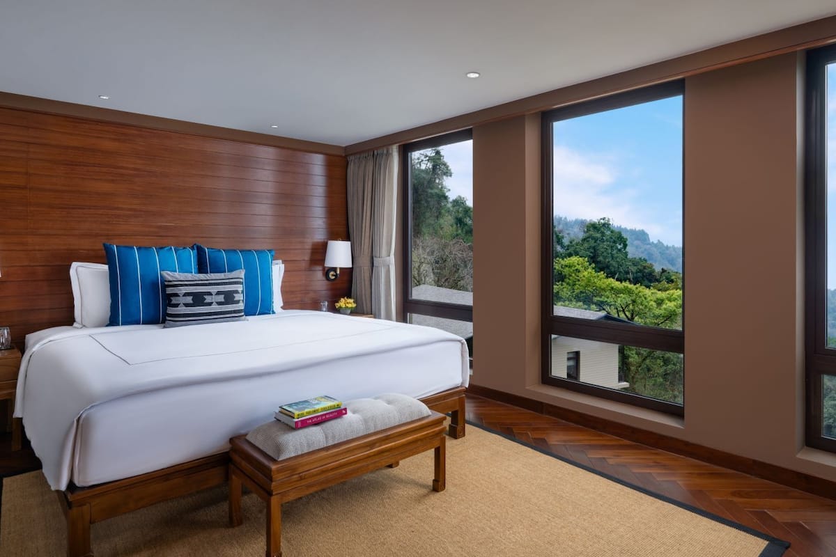 Hotelkamer met een groot bed, houten lambrisering, een bankje, nachtkastje, lamp en uitzicht op een bergachtig landschap door grote ramen.