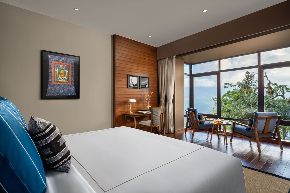 A modern hotel room with a large bed, a desk, two chairs, and a view of the outdoors.