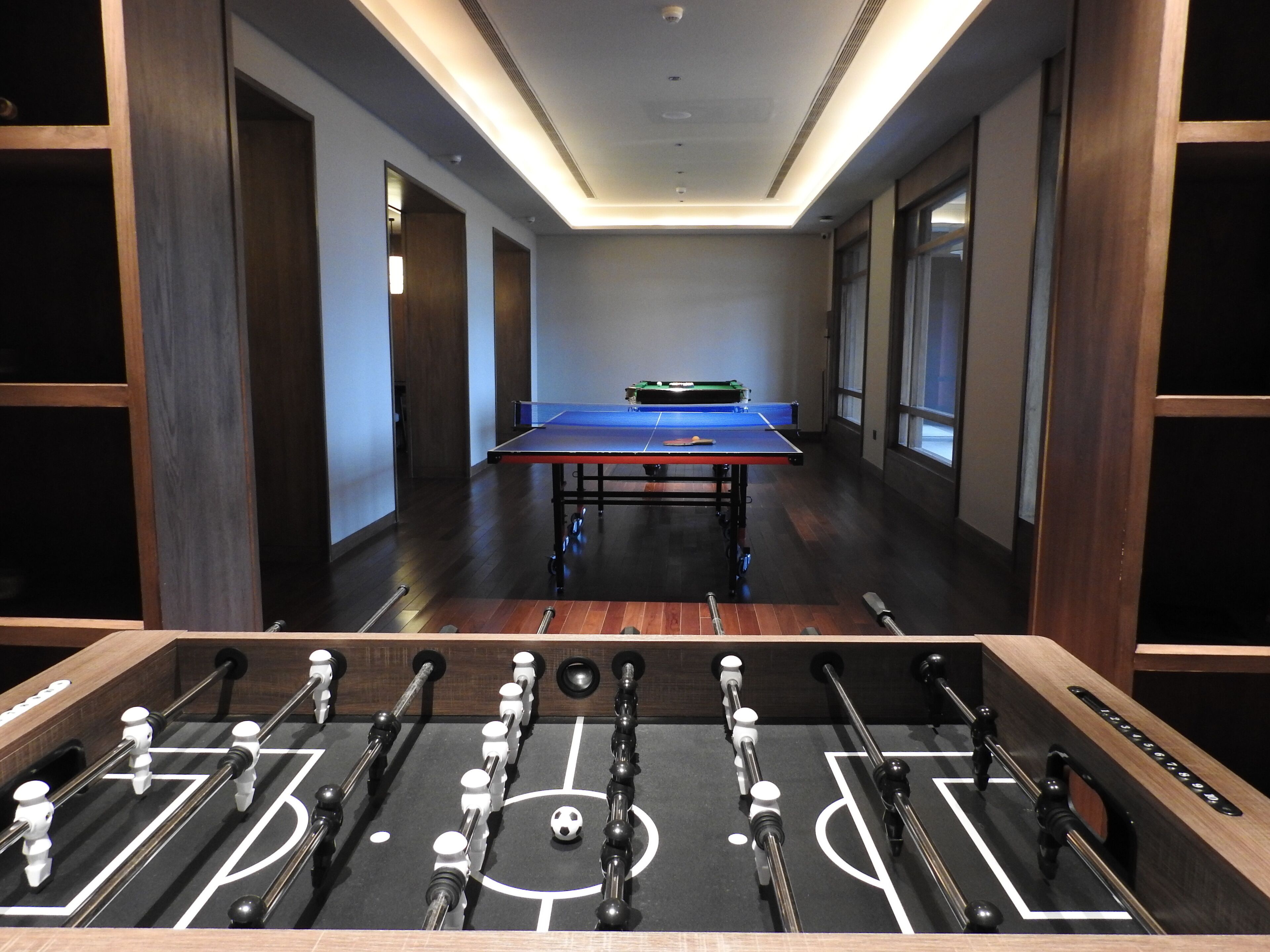A room with a foosball table in the foreground, a ping pong table in the center, and wooden paneling on the walls.